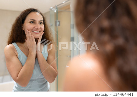 Smiling adult woman touching face while looking in bathroom mirror at home Smiling adult woman touching face while looking in bathroom mirror at home 134464441
