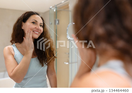 Smiling adult woman touching her cheek while looking in bathroom mirror 134464460