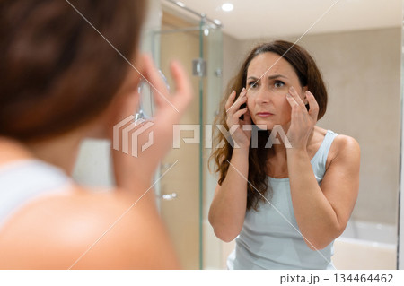 Concerned adult woman examining under eye skin in bathroom mirror Concerned adult woman examining under eye skin in bathroom mirror 134464462