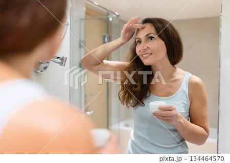 Adult woman applying face cream on forehead and smiling at mirror reflection 134464570