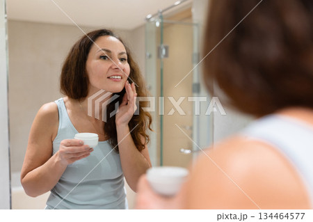 Adult woman applying face cream while looking in bathroom mirror Adult woman applying face cream while looking in bathroom mirror 134464577