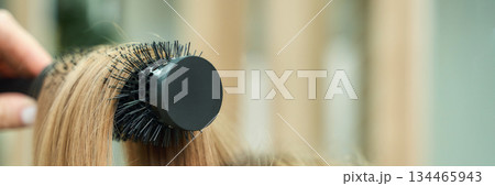 Close-up of round hairbrush styling blonde hair with hand indoors 134465943