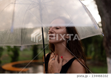 Woman, rain, umbrella. Serene woman enjoying a peaceful moment under a clear umbrella outdoors. 134466102