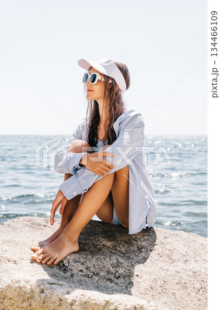 Woman Ocean Relaxing: Barefoot woman sits on coastal rock, enjoying sunny summer day by blue sea. 134466109