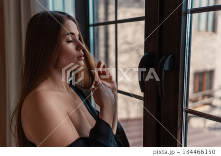 Woman, window, morning light illuminates young female touching long brown hair in sensual moment Woman, window, morning light illuminates young female touching long brown hair in sensual moment 134466150
