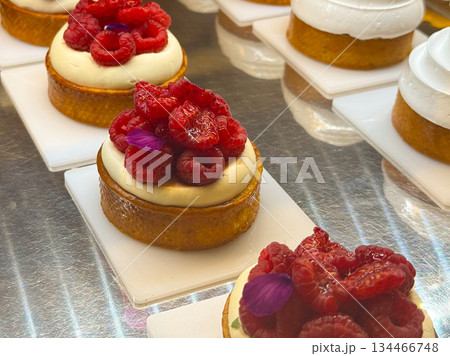 Round pastries with white meringue topping and cakes decorated with red berries arranged on white plates. Bakery display combining cream, color and craft. 134466748