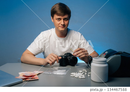 Cocaine dealer sitting at table, displaying drug paraphernalia, cash and pills, holding rolled banknote, looking at camera, studio shot portrait on isolated background. Concept of substance abuse. Cocaine dealer sitting at table, displaying drug paraphernalia, cash and pills, holding rolled banknote, looking at camera, studio shot portrait on isolated background. Concept of substance abuse. 134467588