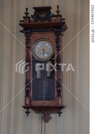 Antique wooden wall clock with carved details, pendulum and weights mounted on striped wallpaper, warm brown tones and vintage craftsmanship Antique wooden wall clock with carved details, pendulum and weights mounted on striped wallpaper, warm brown tones and vintage craftsmanship 134467602