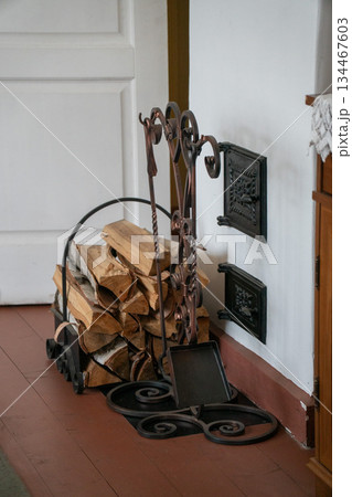 Wrought iron fireplace tool set with stacked firewood stands beside a traditional stove inside a cozy interior, warm tones and rustic details Wrought iron fireplace tool set with stacked firewood stands beside a traditional stove inside a cozy interior, warm tones and rustic details 134467603