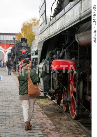 Woman in a green jacket stands beside a vintage steam locomotive with red wheels on an outdoor railway platform, industrial details and autumn mood 134467605