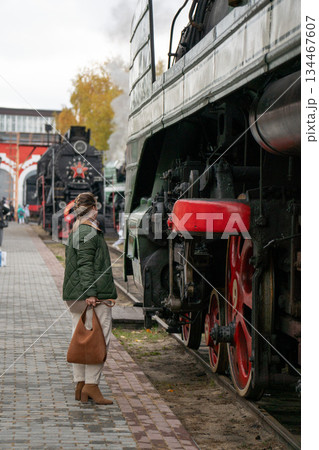 Woman in a green jacket stands beside a vintage steam locomotive with red wheels on an outdoor railway platform, industrial details and autumn mood Woman in a green jacket stands beside a vintage steam locomotive with red wheels on an outdoor railway platform, industrial details and autumn mood 134467607