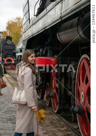 Woman stands beside preserved Soviet-era steam locomotive, looking at large red driving wheels at the Povorotny Krug railway museum 134467611