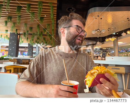 A man with glasses enjoys a taco and drink while sitting inside a food court restaurant. Casual dining, consumer lifestyle, fast food culture and modern gastronomy in everyday leisure. A man with glasses enjoys a taco and drink while sitting inside a food court restaurant. Casual dining, consumer lifestyle, fast food culture and modern gastronomy in everyday leisure. 134467773