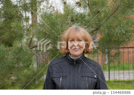 Portrait of a happy elderly beautiful woman near beautiful fir trees in her garden. Portrait of a happy elderly beautiful woman near beautiful fir trees in her garden. 134468986