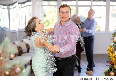 Active middle-aged man and mature woman in pair practicing ballroom dance in training hall during Christmas party 134470930