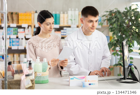 Guy pharmacist at counter with computer sells pills, consulting girl about doctors prescription. 134471346