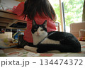部屋の中で女の子の後ろに寄り添って座る猫 134474372