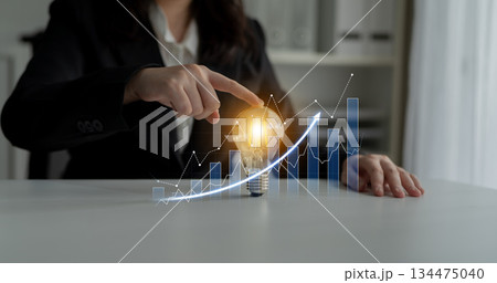 Businesswoman in Suit Pointing at Light Bulb with Rising Graphs, Illustrating Innovation and Growth in Corporate Strategy and Decision-Making Process Scalp 134475040
