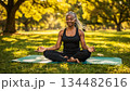 elderly African American woman sits on a mat in the lotus position in a sunny meadow in the morning park, practicing yoga for health and longevity 134482616