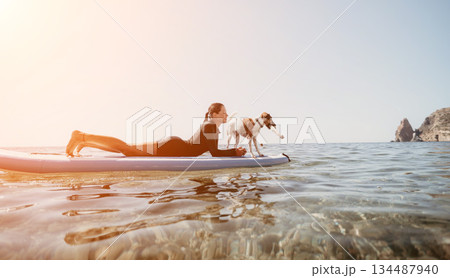 Woman Dog Paddleboard Sunset - Woman and dog paddleboarding together on a calm ocean at sunset. 134487940