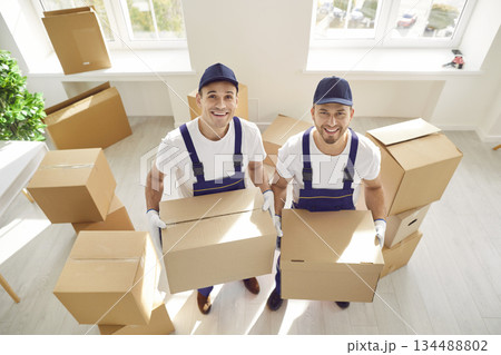 Portrait of two smiling professional movers effortlessly carrying cardboard boxes into room of house Portrait of two smiling professional movers effortlessly carrying cardboard boxes into room of house 134488802