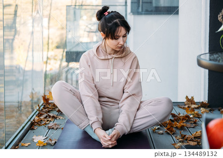 Woman performs butterfly stretch on yoga mat amidst fallen leaves on balcony. Urban living, private sanctuary, home fitness. Woman performs butterfly stretch on yoga mat amidst fallen leaves on balcony. Urban living, private sanctuary, home fitness. 134489132