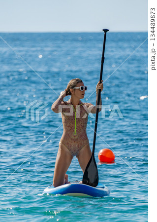 Paddleboarding, woman, ocean. Young woman kneeling on stand-up paddleboard in blue sea enjoying summer water sport activity. Paddleboarding, woman, ocean. Young woman kneeling on stand-up paddleboard in blue sea enjoying summer water sport activity. 134489873