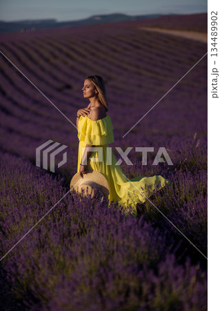 Blonde woman standing in vast purple lavender field holding straw hat at sunset Blonde woman standing in vast purple lavender field holding straw hat at sunset 134489902