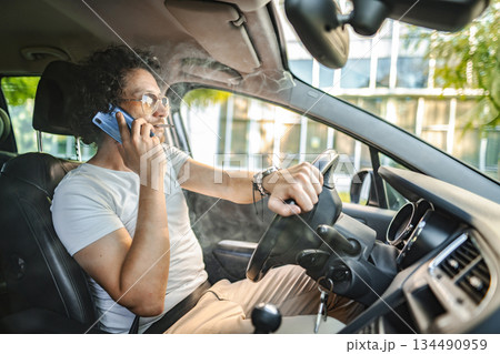 Man with curly hair driving a car, smoking a cigarette and using phone 134490959