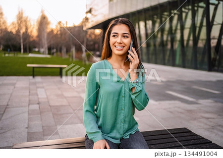 Young businesswoman talking on smartphone while sitting on bench in urban park 134491004