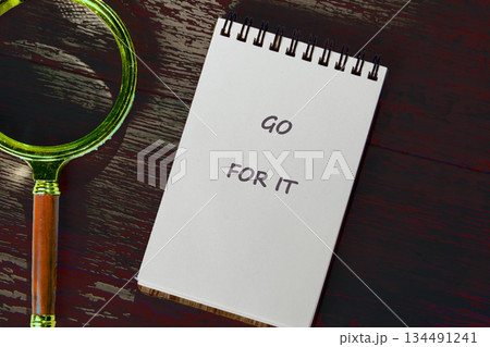 A notepad displaying GO FOR IT rests near a golden magnifying glass on a weathered wood surface. This scene inspires motivation and exploration of possibilities 134491241