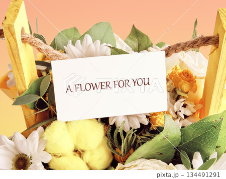 A floral gift basked in glowing colors. Daisies and roses sit pretty in a wooden basket with a card that says A FLOWER FOR YOU, perfect for sharing 134491291
