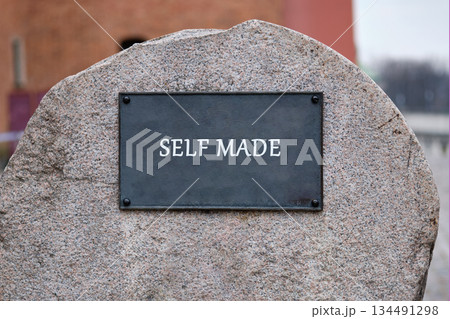 A stone monument displays a plaque with the phrase SELF MADE in a simple typeface. The area around the monument is blurry, suggesting it is located in a city 134491298