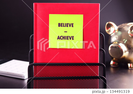 A bright yellow sticky note with the phrase BELIEVE - ACHIEVE is adhered to a vibrant red folder. The folder sits neatly in a black metal mesh organizer on a dark desktop 134491319