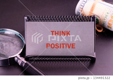 A card reading THINK POSITIVE rests alongside a magnifying glass and a roll of money, suggesting a focus on optimism in financial pursuits A card reading THINK POSITIVE rests alongside a magnifying glass and a roll of money, suggesting a focus on optimism in financial pursuits 134491322
