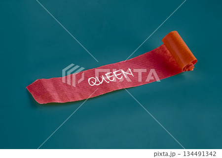 A vibrant, coral-hued paper, scripted with the word Queen in white, casually unrolls across a serene sea-green backdrop, presenting a regal yet lighthearted message A vibrant, coral-hued paper, scripted with the word Queen in white, casually unrolls across a serene sea-green backdrop, presenting a regal yet lighthearted message 134491342
