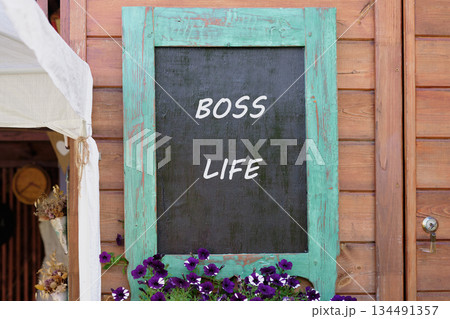 A vintage blackboard displays the words Boss Life in white. Purple flowers flourish below, and the rustic wooden wall adds warmth. It evokes a sense of carefree style A vintage blackboard displays the words Boss Life in white. Purple flowers flourish below, and the rustic wooden wall adds warmth. It evokes a sense of carefree style 134491357