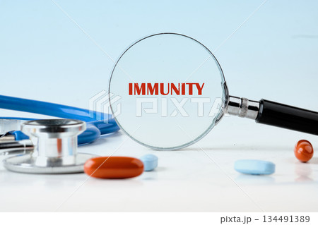 Magnifying glass focuses on the word Immunity, shown in red text. Stethoscope and pills surround the glass, against a light blue backdrop representing healthcare research 134491389