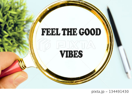 A hand holds a golden magnifying glass showing the words FEEL THE GOOD VIBES near a small plant and a black pen, radiating optimism and hopeful sentiments 134491430