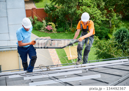 Workers building solar panel system on roof of house. Two men installers in helmets carrying photovoltaic solar module outdoors. Alternative, green and renewable energy generation concept. Workers building solar panel system on roof of house. Two men installers in helmets carrying photovoltaic solar module outdoors. Alternative, green and renewable energy generation concept. 134492367
