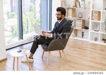 An Indian businessman is working on his laptop while sitting in a modern office setting An Indian businessman is working on his laptop while sitting in a modern office setting 134494165