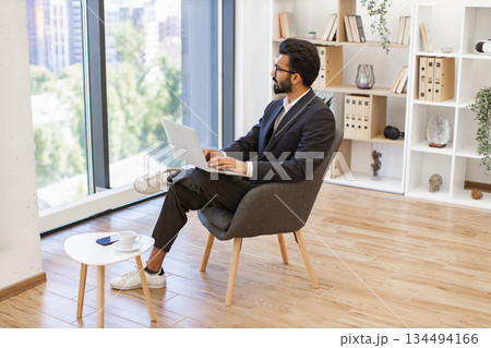 An Arab businessman is working on his laptop while sitting in a modern office, looking out the window An Arab businessman is working on his laptop while sitting in a modern office, looking out the window 134494166