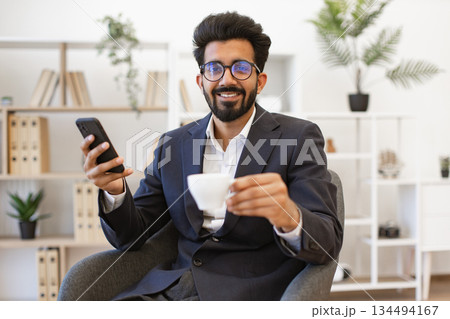 An Indian businessman in a suit smiles while holding a cup of coffee and a smartphone An Indian businessman in a suit smiles while holding a cup of coffee and a smartphone 134494167