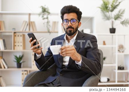An Arab businessman in a suit is holding a cup of coffee and a smartphone while looking at the camera An Arab businessman in a suit is holding a cup of coffee and a smartphone while looking at the camera 134494168