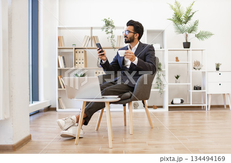 An Arab businessman is relaxing in a modern office setting, holding a phone and a cup of coffee 134494169