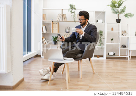 A stylish Arab businessman is relaxing in a modern office setting, using his phone and enjoying a coffee break A stylish Arab businessman is relaxing in a modern office setting, using his phone and enjoying a coffee break 134494533