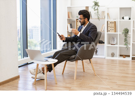 An Indian businessman is relaxing in a modern office while holding a cup of coffee and looking at his phone 134494539