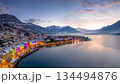 Domaso Lake Como Italy winter cityscape at sunset with colorful rainbow Christmas lights on waterfront buildings, snow covered mountains, misty alpine lake reflections and scenic twilight sky. 134494876
