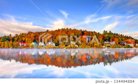 Scenic autumn landscape of Saranac Lake village in Adirondack Mountains New York featuring colorful fall foliage, historic waterfront houses and perfect misty water reflection under blue sky. 134494884