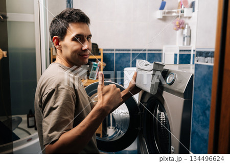 Portrait of man showing thumbs up gesture looking camera, holding lint filter, removing from tumble dryer in laundry room, performing appliance cleaning and maintenance to ensure proper function. Portrait of man showing thumbs up gesture looking camera, holding lint filter, removing from tumble dryer in laundry room, performing appliance cleaning and maintenance to ensure proper function. 134496624